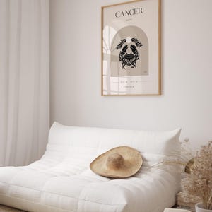 May include: A minimalist poster with a beige background and a black crab illustration. The poster has the text "CANCER MOON" and "JUNE 21 - JULY 22" in black text. The poster is framed in a light wood frame and is hanging on a white wall.