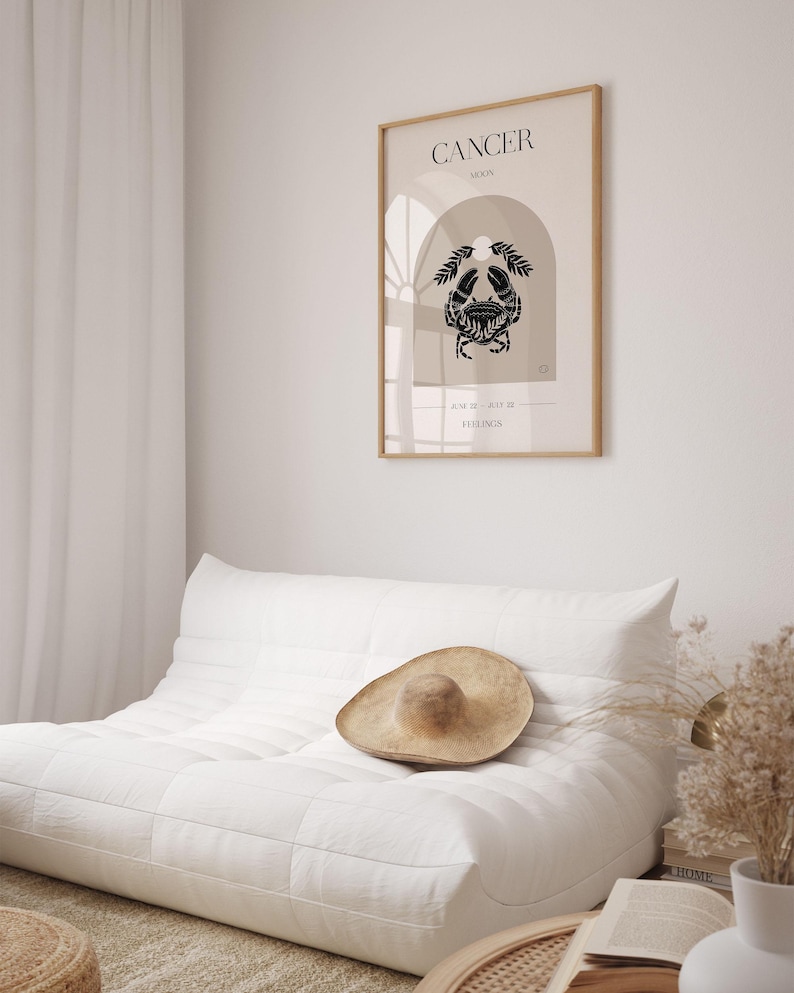 May include: A minimalist poster with a beige background and a black crab illustration. The poster has the text "CANCER MOON" and "JUNE 21 - JULY 22" in black text. The poster is framed in a light wood frame and is hanging on a white wall.