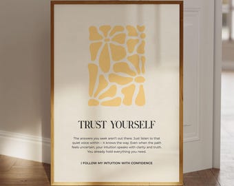 May include: A framed art print with a cream-colored background and a gold-colored wooden frame. The print features a yellow floral design and the words "TRUST YOURSELF" in black text. Below the text is a paragraph of text and the phrase "I FOLLOW MY INTUITION WITH CONFIDENCE."