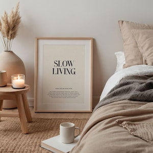 May include: A framed print with the text "Slow Living" in black lettering on a cream background. The print is hanging on a white wall above a wooden stool with a candle on it. The print is in a bedroom setting with a bed in the background.