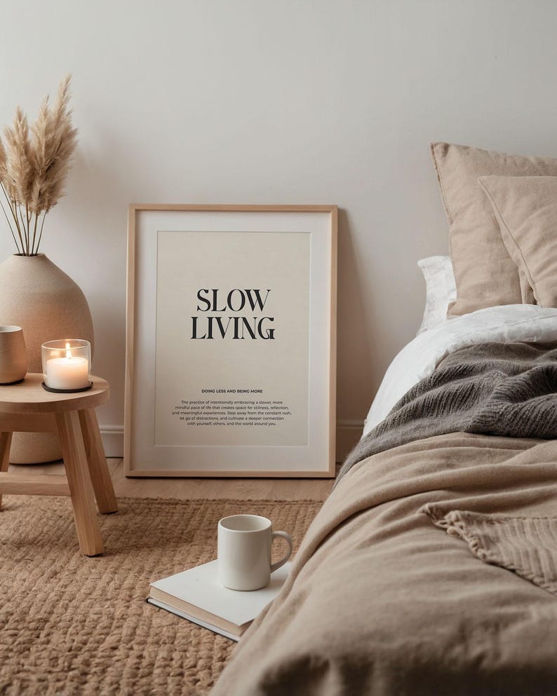 May include: A framed print with the text "Slow Living" in black lettering on a cream background. The print is hanging on a white wall above a wooden stool with a candle on it. The print is in a bedroom setting with a bed in the background.
