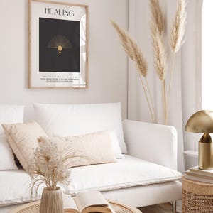 May include: A framed print with the word "HEALING" in black text on a black background. The print features a gold geometric design with a sun and a circle. The print is hanging on a white wall above a white couch with a beige throw pillow. A vase with dried flowers sits on a woven coffee table in front of the couch.