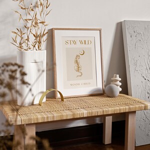 May include: A framed print with a beige background and gold text that reads "Stay Wild" and "Moon Child". A gold outline of a tiger sitting and looking up at a crescent moon is in the center of the print. The print is sitting on a light brown wicker bench.