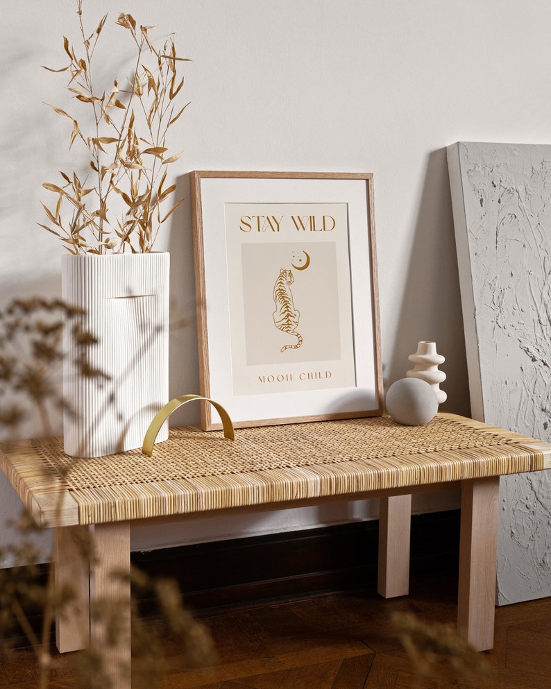 May include: A framed print with a beige background and gold text that reads "Stay Wild" and "Moon Child". A gold outline of a tiger sitting and looking up at a crescent moon is in the center of the print. The print is sitting on a light brown wicker bench.