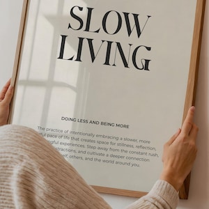 May include: A framed print with the text "SLOW LIVING" in black font on a white background. The text "DOING LESS AND BEING MORE" is below the main text. The print is framed in a light wood frame.