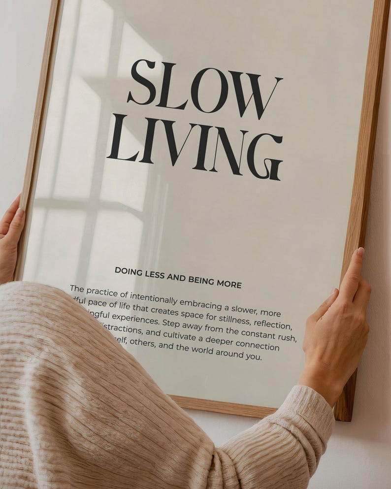 May include: A framed print with the text "SLOW LIVING" in black font on a white background. The text "DOING LESS AND BEING MORE" is below the main text. The print is framed in a light wood frame.