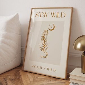 May include: A beige poster with a gold outline and the text "STAY WILD" and "MOON CHILD" in a gold script font. A gold silhouette of a tiger sitting and looking up at a crescent moon with stars is in the center of the poster.