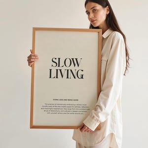 May include: A framed print with the text "SLOW LIVING" in black lettering on a white background. The text "DOING LESS AND BEING MORE" is below the main text in a smaller font. The print is framed in a light brown wood frame.