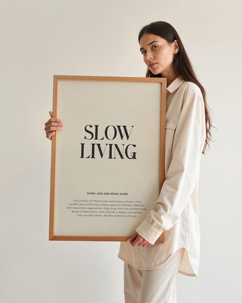 May include: A framed print with the text "SLOW LIVING" in black lettering on a white background. The text "DOING LESS AND BEING MORE" is below the main text in a smaller font. The print is framed in a light brown wood frame.
