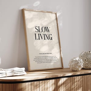 May include: A framed print with the text "SLOW LIVING" in black lettering on a beige background. The text "DOING LESS AND BEING MORE" is below the main text, followed by a short paragraph about the practice of slow living.