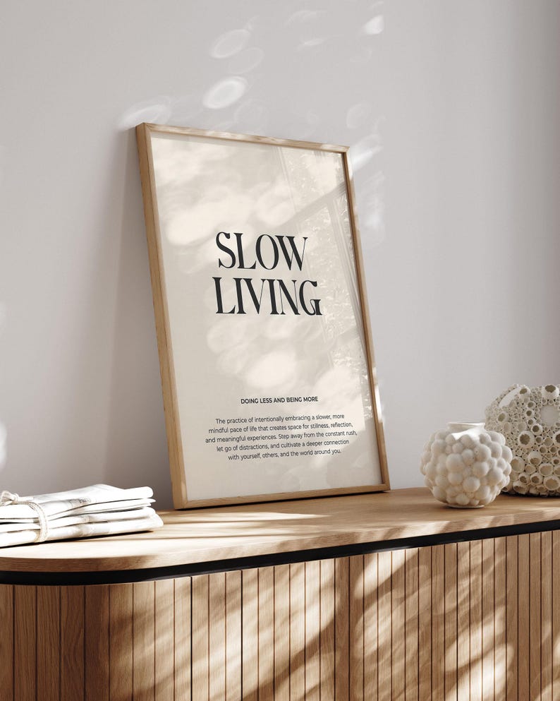 May include: A framed print with the text "SLOW LIVING" in black lettering on a beige background. The text "DOING LESS AND BEING MORE" is below the main text, followed by a short paragraph about the practice of slow living.