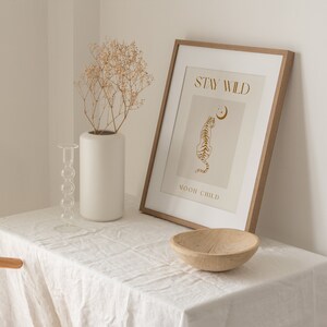 May include: A framed print with a beige background and gold text that reads "Stay Wild" and "Moon Child". A gold outline of a tiger is facing left with a crescent moon above it. The frame is light brown wood.