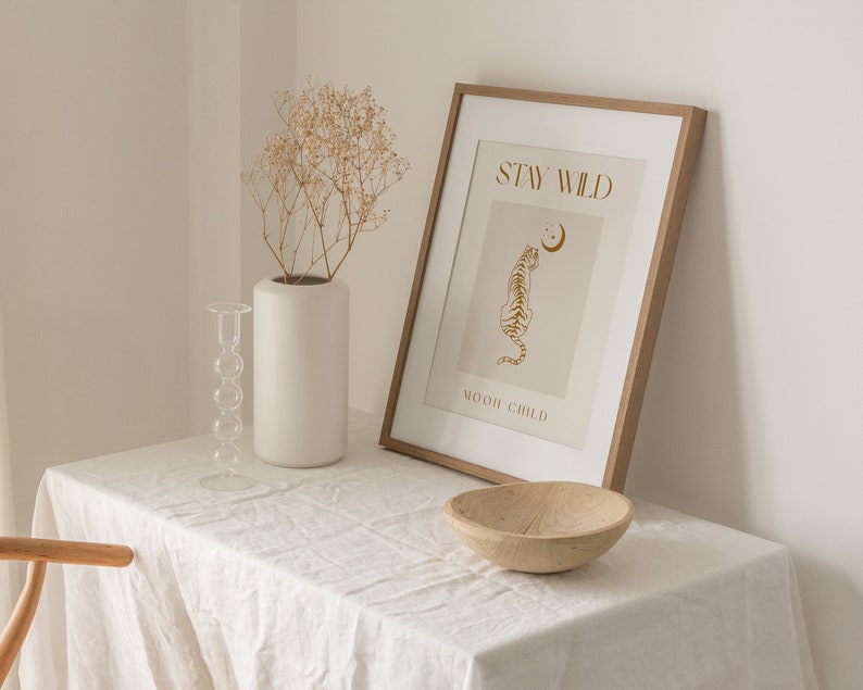 May include: A framed print with a beige background and gold text that reads "Stay Wild" and "Moon Child". A gold outline of a tiger is facing left with a crescent moon above it. The frame is light brown wood.