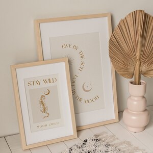 May include: Two framed prints with inspirational quotes and illustrations. The top print says "Live by the sun, love by the moon." The bottom print says "Stay Wild" and features a tiger illustration. The prints are in light wood frames and are displayed on a white surface.