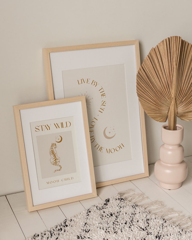 May include: Two framed prints with inspirational quotes and illustrations. The top print says "Live by the sun, love by the moon." The bottom print says "Stay Wild" and features a tiger illustration. The prints are in light wood frames and are displayed on a white surface.
