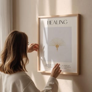 May include: A framed print with the word "HEALING" in black text at the top. The print features a stylized sunburst design in gold with a white background. The text below the design reads "Healing is the art of turning shadows into love and knowing peace with yourself and others. Return to unconditional love and be ready whole."