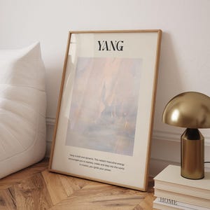 Op de afbeelding: Een ingelijste kunstafdruk met het woord "YANG" bovenaan. Het kunstwerk heeft een abstract ontwerp in tinten beige, blauw en oranje. De afdruk leunt tegen een witte muur en een houten vloer. Een gouden lamp en boeken zijn ook zichtbaar.