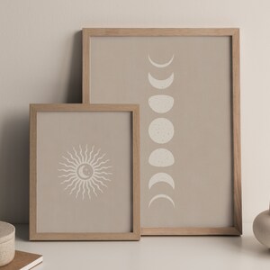 May include: Two framed prints with a beige background. The print on the left features a stylized sun with rays. The print on the right features a series of crescent moons in different phases.