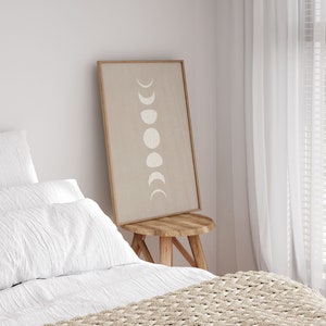 May include: A framed print of a beige background with white crescent moons in various phases. The print is hanging on a white wall above a wooden stool. The stool is in front of a bed with white bedding and a beige knitted throw blanket.