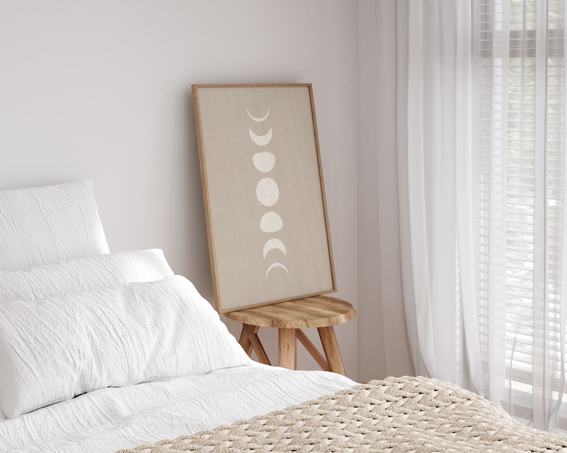 May include: A framed print of a beige background with white crescent moons in various phases. The print is hanging on a white wall above a wooden stool. The stool is in front of a bed with white bedding and a beige knitted throw blanket.