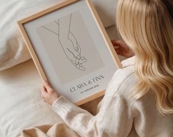 May include: A framed minimalist line art print of two hands holding each other. The print is on a beige background with the text "CLARA & FINN 23. JANUAR 2018" in black.