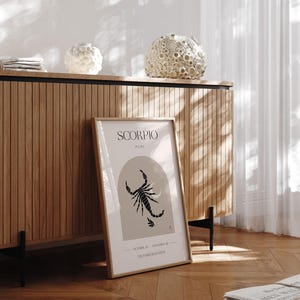 Puede incluir: Una impresi&oacute;n enmarcada con una ilustraci&oacute;n de escorpi&oacute;n negro y el texto "SCORPIO, PLUTO, 23 DE OCTUBRE - 21 DE NOVIEMBRE, TRANSFORMACI&Oacute;N". La impresi&oacute;n est&aacute; sobre un fondo marr&oacute;n claro y est&aacute; enmarcada en un marco marr&oacute;n claro. La impresi&oacute;n est&aacute; colocada sobre un gabinete de madera con una base de metal negro.