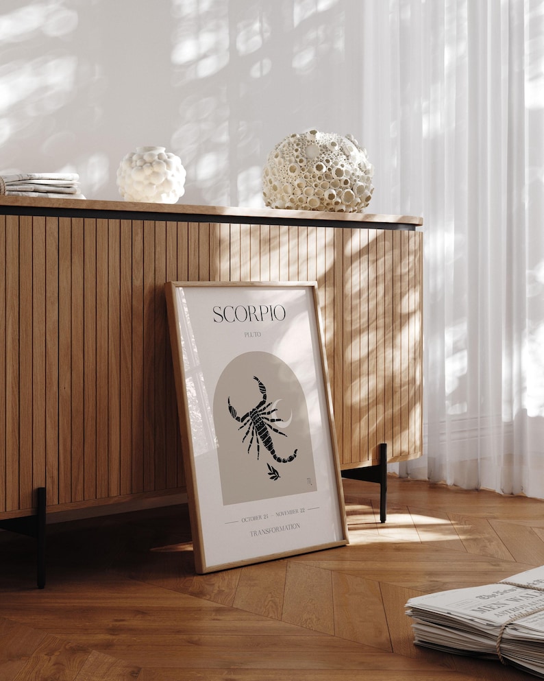 Puede incluir: Una impresi&oacute;n enmarcada con una ilustraci&oacute;n de escorpi&oacute;n negro y el texto "SCORPIO, PLUTO, 23 DE OCTUBRE - 21 DE NOVIEMBRE, TRANSFORMACI&Oacute;N". La impresi&oacute;n est&aacute; sobre un fondo marr&oacute;n claro y est&aacute; enmarcada en un marco marr&oacute;n claro. La impresi&oacute;n est&aacute; colocada sobre un gabinete de madera con una base de metal negro.