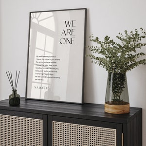 May include: A black framed poster with a white background and the text "WE ARE ONE" in large, bold letters. The poster also includes a poem about honoring the soul and the universe within each person. The poster is hanging on a wall above a black cabinet with a woven rattan front.