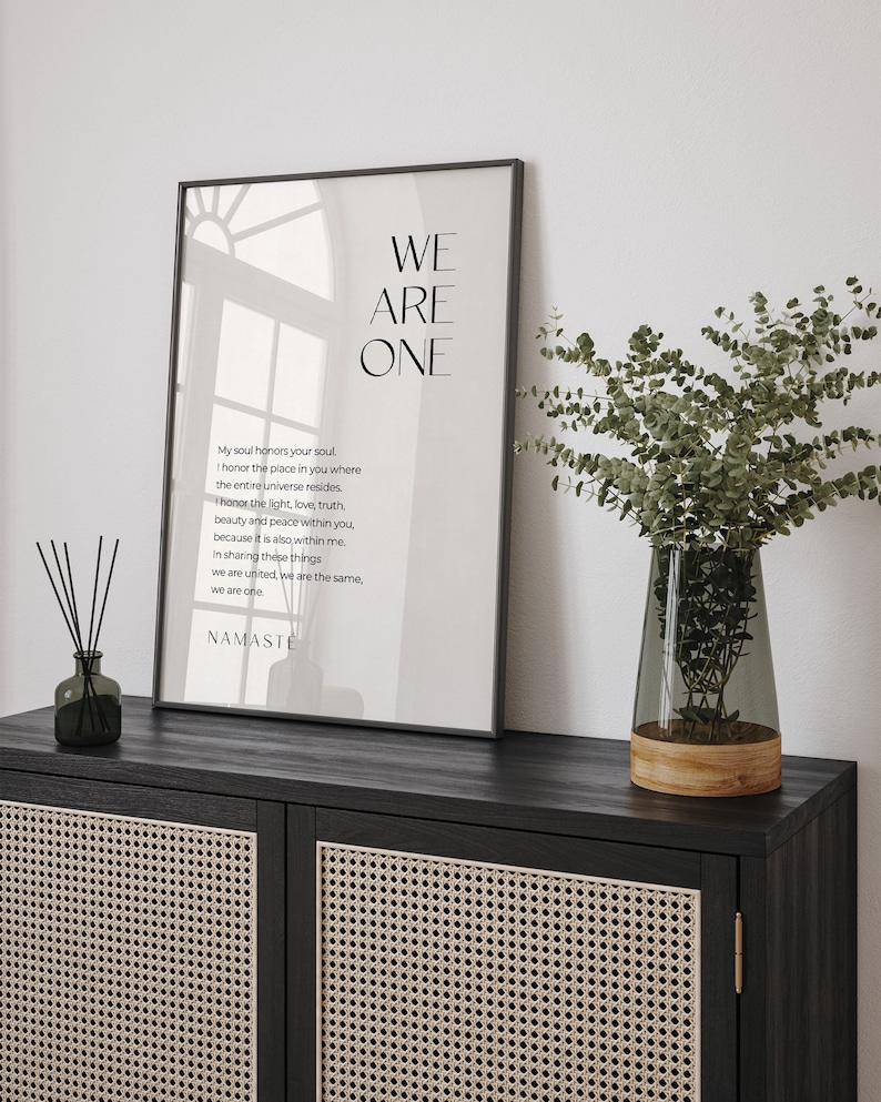 May include: A black framed poster with a white background and the text "WE ARE ONE" in large, bold letters. The poster also includes a poem about honoring the soul and the universe within each person. The poster is hanging on a wall above a black cabinet with a woven rattan front.