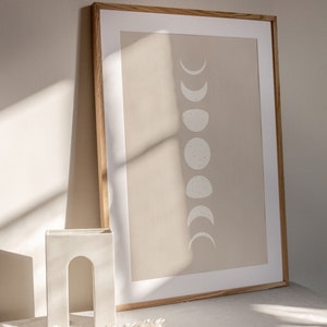 May include: A framed print with a beige background and a white silhouette of the moon in various phases. The frame is made of wood and has a light brown finish. The print is sitting on a white table with a white ceramic arch-shaped vase and dried flowers.
