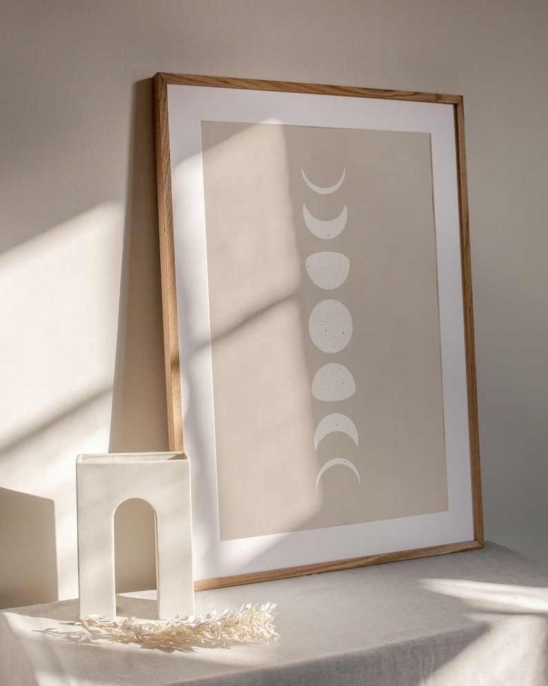 May include: A framed print with a beige background and a white silhouette of the moon in various phases. The frame is made of wood and has a light brown finish. The print is sitting on a white table with a white ceramic arch-shaped vase and dried flowers.