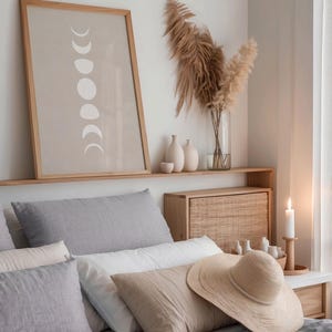 May include: A bedroom with a gray bedspread and a variety of pillows. A framed print of the moon phases hangs above the bed. A straw hat sits on the bed.