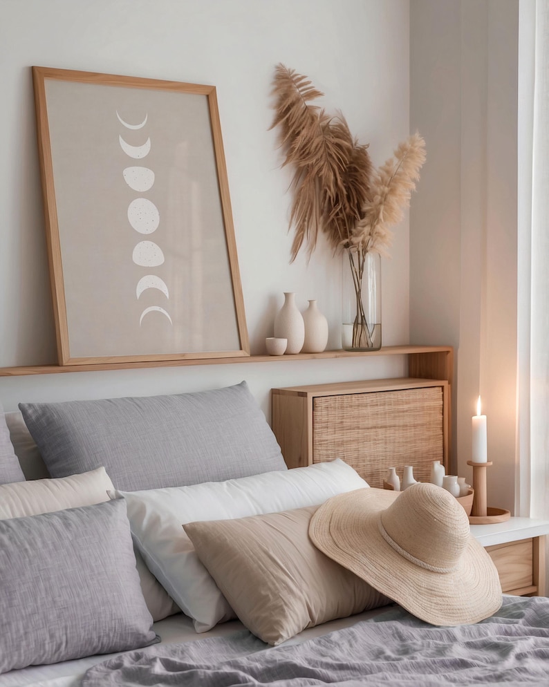 May include: A bedroom with a gray bedspread and a variety of pillows. A framed print of the moon phases hangs above the bed. A straw hat sits on the bed.