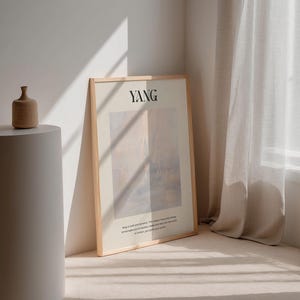 Op de afbeelding: Een ingelijste kunstprint met het woord "YANG" in zwarte letters. Het kunstwerk heeft een zacht, abstract ontwerp in gedempte tinten beige, roze en blauw. Een kleine, bruine keramische vaas staat op een witte sokkel. De print leunt tegen een muur.