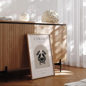 May include: A framed print with a beige background and black illustration of a crab. The text "CANCER MOON" and "JUNE 21 - JULY 22" is printed below the crab. The print is on a wooden surface.