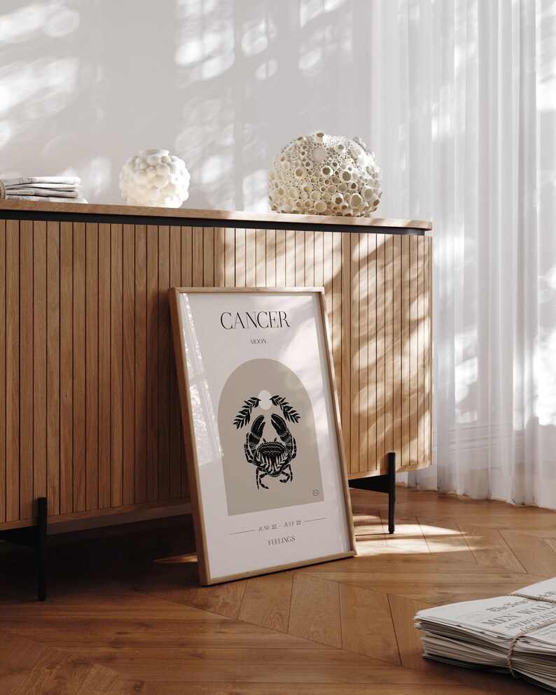 May include: A framed print with a beige background and black illustration of a crab. The text "CANCER MOON" and "JUNE 21 - JULY 22" is printed below the crab. The print is on a wooden surface.