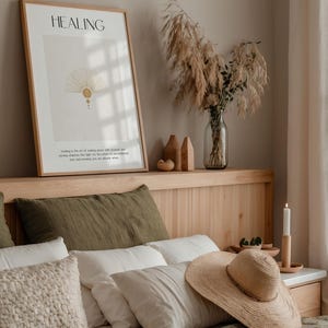 May include: A bedroom with a wooden headboard and a bed with green and white bedding. A framed print with the word "HEALING" and a quote about making peace with yourself is hanging on the wall above the headboard. A vase with dried pampas grass is on the headboard shelf. A straw hat is on the bed.