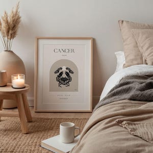 May include: A minimalist poster with a beige background and a black crab illustration. The poster has the text "CANCER MOON" and "AND ALL THE FEELINGS" in black text. The poster is framed in a light wood frame and is hanging on a wall.