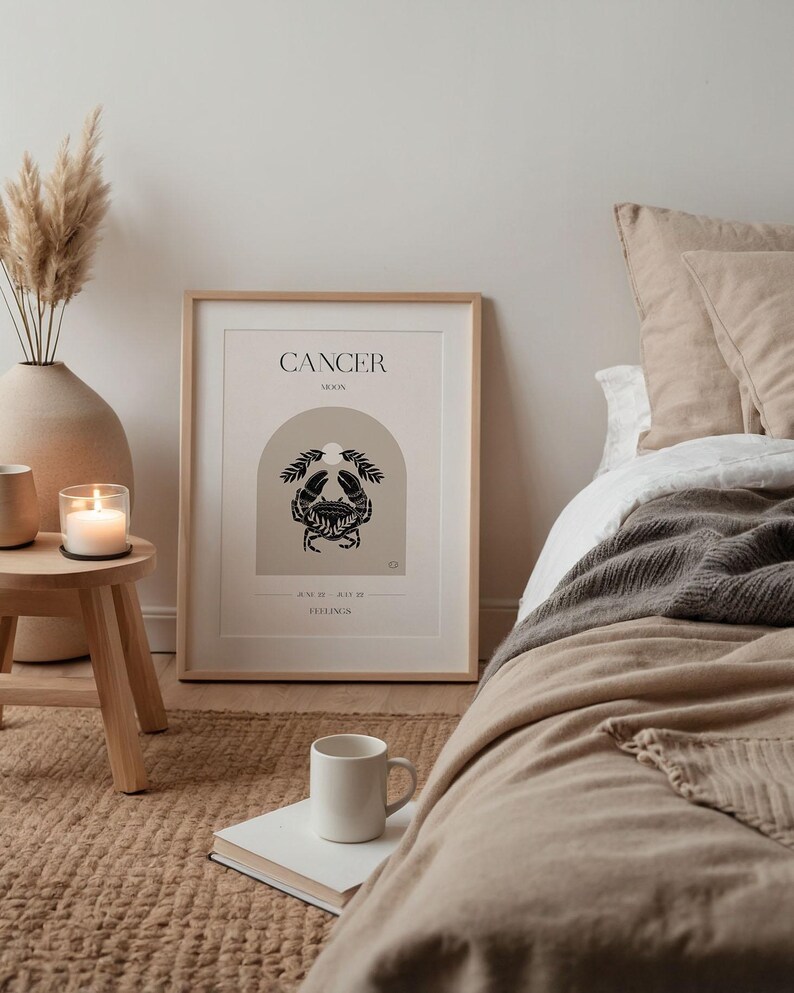 May include: A minimalist poster with a beige background and a black crab illustration. The poster has the text "CANCER MOON" and "AND ALL THE FEELINGS" in black text. The poster is framed in a light wood frame and is hanging on a wall.