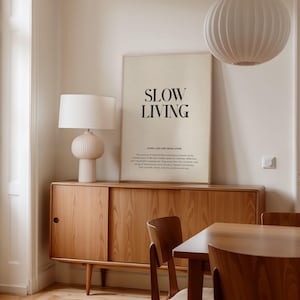 May include: A minimalist poster with the text "SLOW LIVING" in black font on a beige background. The poster is hanging on a white wall above a wooden cabinet with sliding doors. The cabinet is next to a wooden dining table with two chairs. There is a white lampshade hanging from the ceiling and a white table lamp on the cabinet.