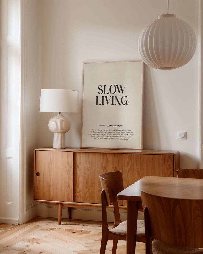 May include: A minimalist poster with the text "SLOW LIVING" in black font on a beige background. The poster is hanging on a white wall above a wooden cabinet with sliding doors. The cabinet is next to a wooden dining table with two chairs. There is a white lampshade hanging from the ceiling and a white table lamp on the cabinet.