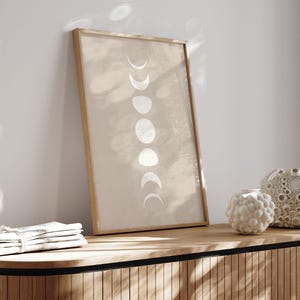 May include: A framed print with a beige background and a vertical row of white crescent moons in various phases. The print is hanging on a light wood wall shelf with a decorative vase on the shelf.