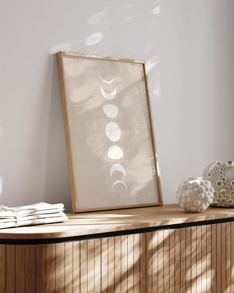 May include: A framed print with a beige background and a vertical row of white crescent moons in various phases. The print is hanging on a light wood wall shelf with a decorative vase on the shelf.