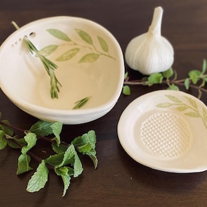May include: White ceramic bowl with green leaf design and a small white ceramic garlic press with a grater surface. Both pieces are on a dark brown wooden surface.