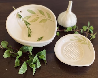 May include: White ceramic bowl with green leaf design and a small white ceramic garlic press with a grater surface. Both pieces are on a dark brown wooden surface.