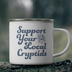 May include: White enamel mug with a silver rim and handle. The mug features the text "Support Your Local Cryptids" in a blue script font, along with a stylized butterfly graphic. The mug is sitting on a dark, textured surface.