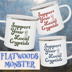 May include: White enamel mugs with silver rims and handles. Each mug features the text "Support Your Local Cryptids" in different colors, along with a small cryptid illustration. The phrase "Flatwoods Monster" is also visible.