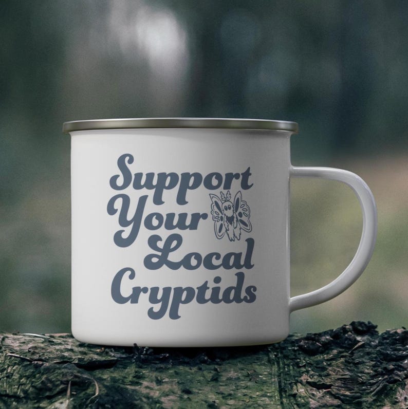 May include: White enamel mug with a silver rim and handle. The mug features the text "Support Your Local Cryptids" in a blue script font, along with a stylized butterfly graphic. The mug is sitting on a dark, textured surface.