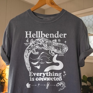 May include: Dark grey t-shirt featuring the word "Hellbender" in white, accompanied by a detailed illustration of a hellbender salamander and aquatic life. The text "Everything is connected" is below the illustration.