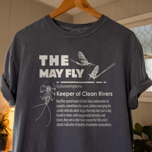 May include: Dark gray t-shirt with the text "THE MAY FLY" and illustrations of mayflies. The shirt also includes the text "Keeper of Clean Rivers" and a description of mayflies.